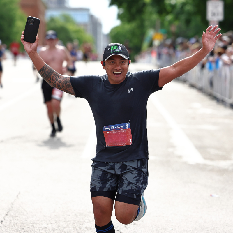 Runner crossing the finish line with arms raised, taking a selfie with a smartphone as cheering spectators watch.