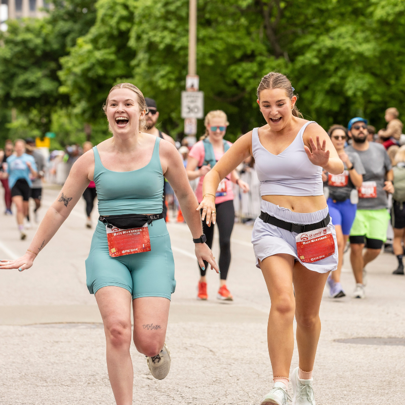 Two women in athletic outfits run toward the camera, smiling and cheering, with spectators in the background during a race on a sunny day.