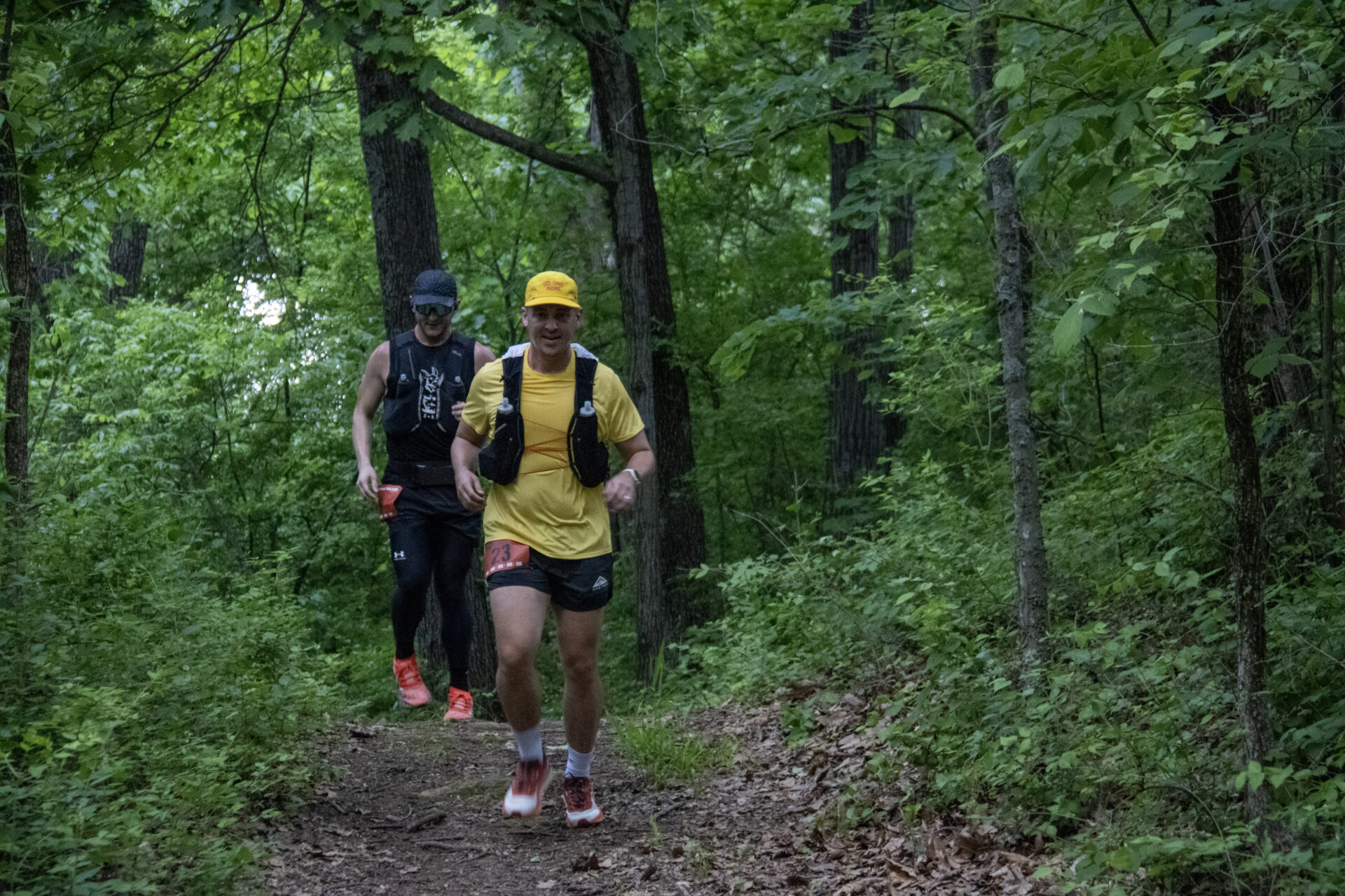 Two runners on a forest trail wearing athletic gear, mid-stride among green trees and foliage.
