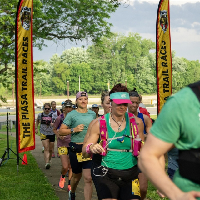 Runners start a trail race under tall yellow PIASA TRAIL RACES banners in a sunny park.