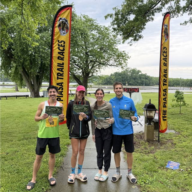 Four runners stand on a park path holding race plaques, with yellow PIASA Trail Races banners and a lake in the background.