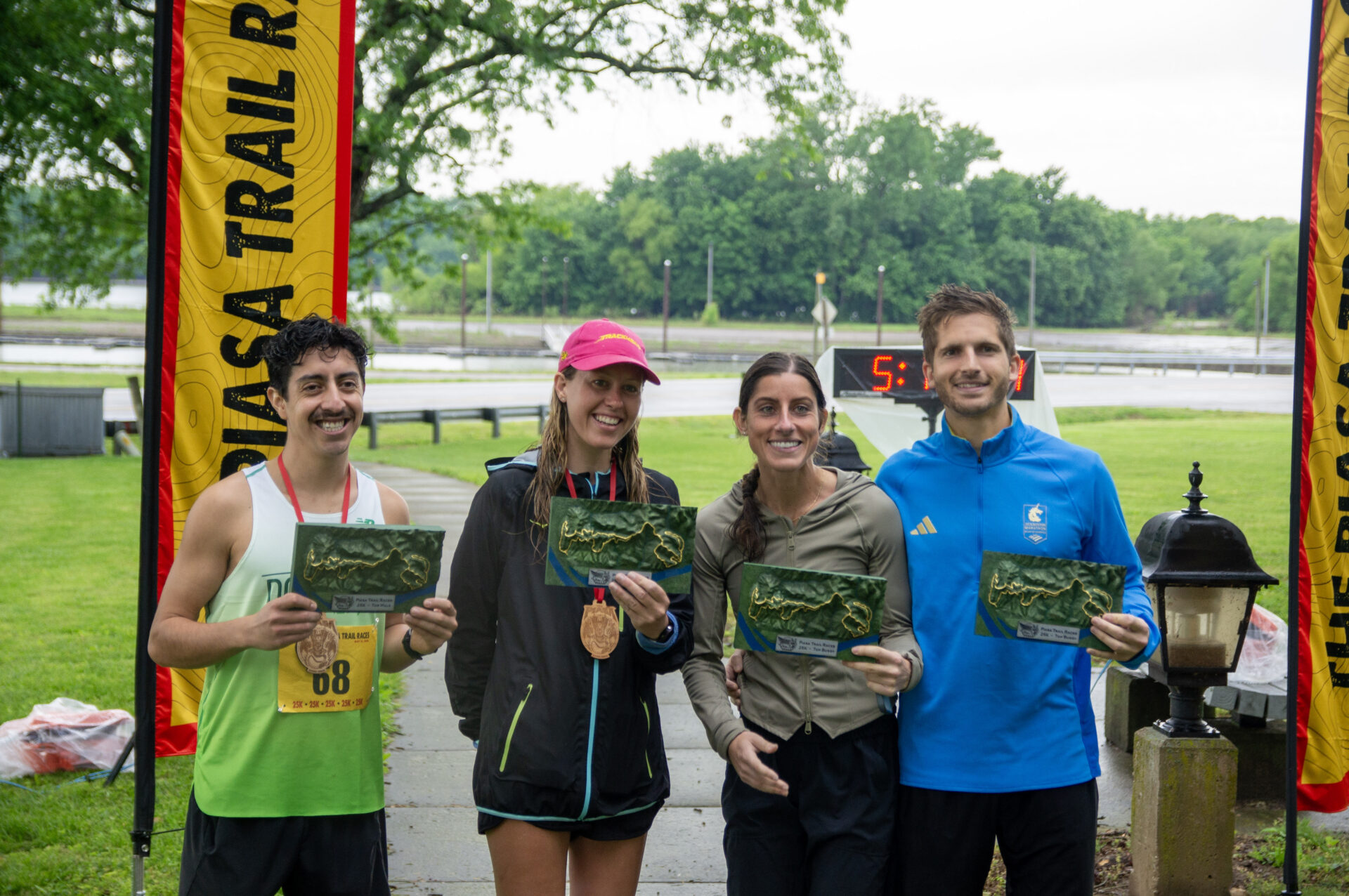 Four smiling runners pose with awards and medals at a race finish line, yellow 'Trail Race' banners in the background, green park setting.
