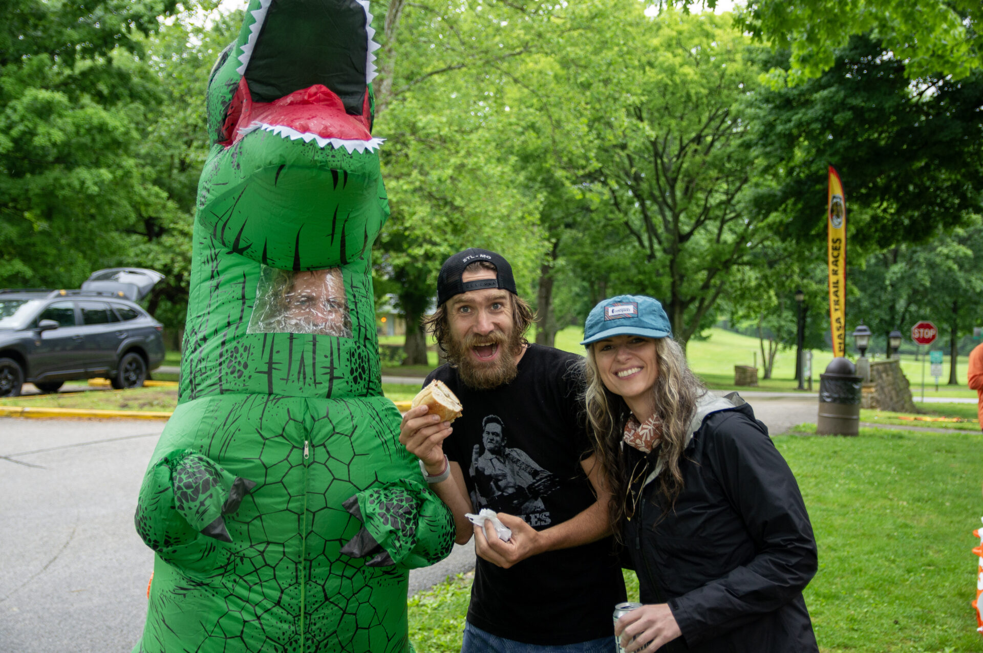 Man and woman smile at the camera next to a person in a large green inflatable dinosaur costume outdoors in a park, holding bread slices.