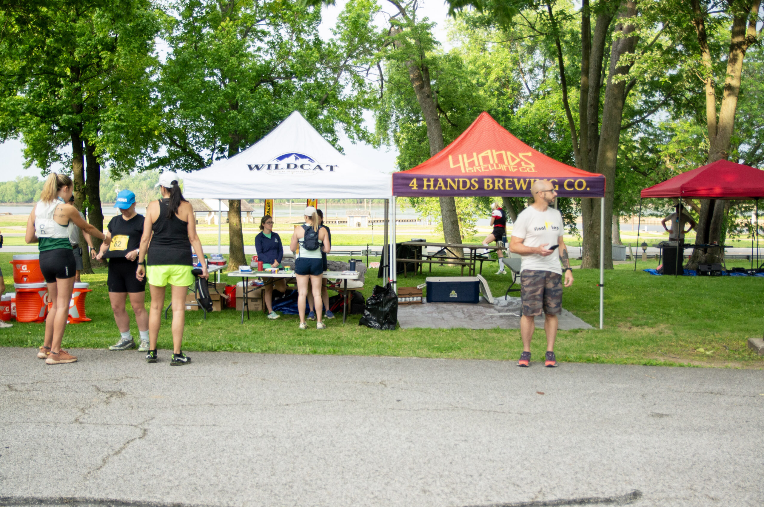 People mingle at an outdoor race event under white and red tents in a sunlit park, with trees in the background and race bibs visible on participants far left.